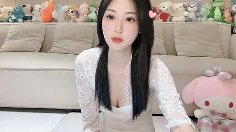 Media: Video of a young East Asian woman with long black hair, fair skin, and a small nose piercing, wearing a white lace dress, sitting in a plush, cozy living room filled with stuffed animals.