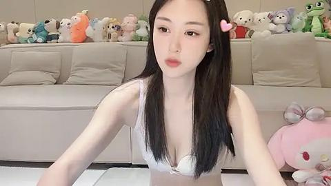 Media: Video of an East Asian woman with long black hair, fair skin, and a petite frame, wearing a white lace bra, sitting on a white carpet in a cozy living room filled with plush toys and a beige couch.