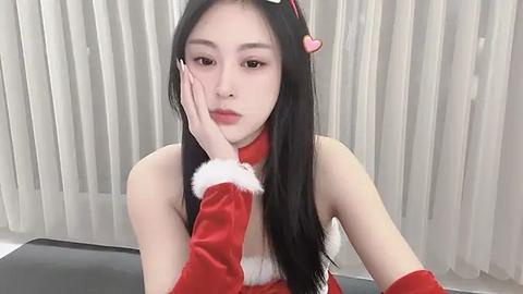 Media: Video of a young East Asian woman with long black hair, wearing a red velvet Santa hat and sleeveless dress, leaning on a hand against a white curtain background.