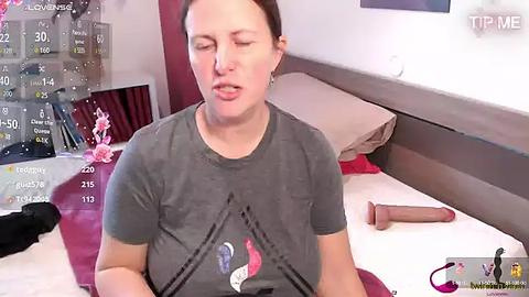 Media: Video of a woman with short brown hair in a gray T-shirt, lying on a bed in a messy bedroom with scattered clothes and a dildo on the bed.