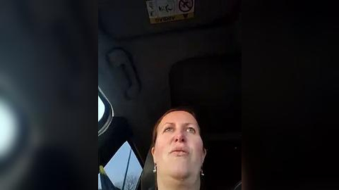 Media: Video of a woman with light skin and brown hair, wearing a black top, looking out a car window. She has a pensive expression. The car interior is dimly lit, with a sign and a seatbelt visible.