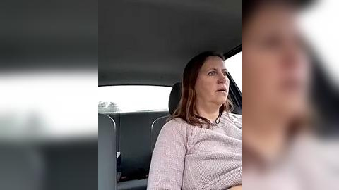 Media: Video of a middle-aged woman with shoulder-length brown hair, wearing a light pink knit sweater, sitting in a car with a blurred, indistinct person in the foreground.