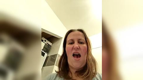 Media: Video of a middle-aged woman with shoulder-length brown hair, wearing a light blue shirt, smiling and opening her mouth wide. Background features a white wall with a built-in shelf holding laundry items.