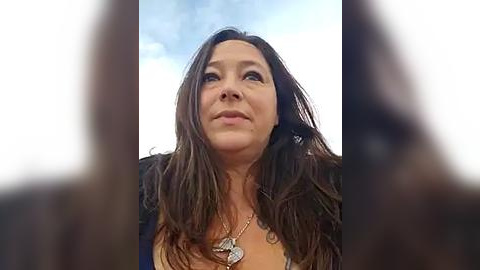 Media: A close-up video of a middle-aged woman with long, wavy brown hair, wearing a necklace and a dark jacket, looking upward with a contemplative expression against a cloudy sky background.