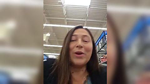 Media: Video of an Asian woman with long, straight brown hair, wearing a black jacket, eyes closed, smiling, standing in a brightly lit, modern grocery store with white ceiling panels and signage.