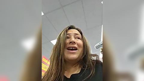 Media: Video of a smiling Asian woman with long, straight, light brown hair, wearing a black shirt, in a brightly lit, modern office with fluorescent lights and a ceiling fan.