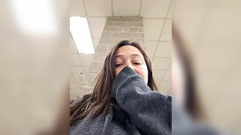 Media: A video of an Asian woman with long, straight brown hair, wearing a gray hoodie, covering her face with her hand, in a fluorescent-lit room with a brick wall and ceiling tiles.