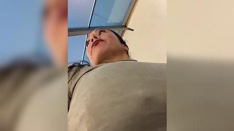 Media: A video of a person looking up at a skylight from a low angle, emphasizing their large, round breasts in a tight, light-colored top. The background is a clear blue sky.