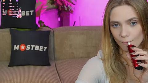 Media: A video of a blonde woman with blue eyes smoking a red cigarette on a beige couch. Behind her, a black pillow with \"MOSTBET\" logo in white and red. Purple ambient lighting.