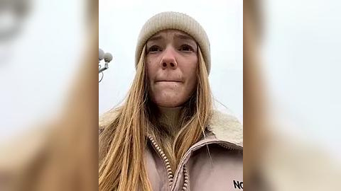 Media: Video of a young Caucasian woman with long, straight blonde hair, wearing a beige knit beanie and a light-colored jacket with a fur-trimmed hood. She has a neutral expression, standing in an outdoor, snowy environment.