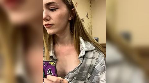 Media: Video of a young woman with straight, light brown hair, wearing a partially unbuttoned plaid shirt, holding a purple Longhorn-themed phone case. The background is a blurred indoor setting with beige walls.