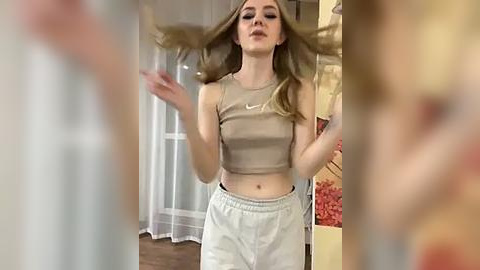 Media: Video of a young Caucasian woman with long blonde hair, wearing a beige crop top and white pants, dancing energetically in a warmly lit room with beige curtains and floral decor.