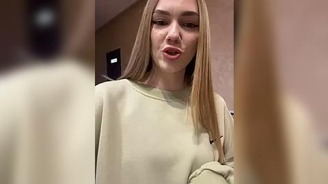 Media: Video of a young woman with long, straight, blonde hair, wearing a cream-colored sweater, puckering her lips in a playful expression. Background features a beige wall and a blurred figure.