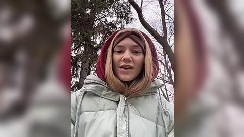 Media: Video of a young girl with light brown skin, wearing a puffy, light green jacket and a red hood, standing outdoors amidst leafless trees in a winter setting.