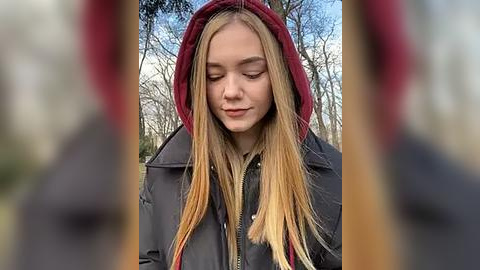 Media: Video of a young woman with long, blonde hair in a dark jacket, hood up, standing in a forest with bare trees and a blue sky.