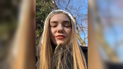 Media: Video of a young, fair-skinned woman with long, straight blonde hair, wearing a white headscarf, standing outdoors against a clear blue sky and leafless trees.