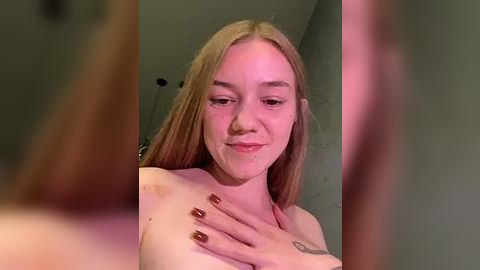 Media: Video of a young Caucasian woman with straight, light brown hair, smiling, topless, with a tattoo on her left shoulder, holding her chest.