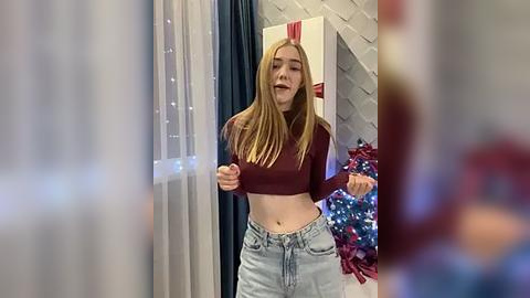 Media: Video of a young woman with long blonde hair, wearing a maroon crop top and light-wash jeans, smiling and holding a pom-pom. Background includes a white gift box with red ribbon, blue and red tinsel, and a curtain.