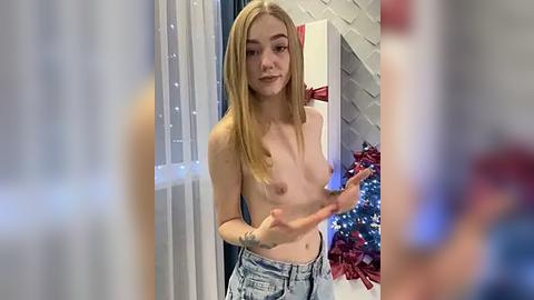 Media: Video of a topless young Caucasian woman with small breasts, blonde hair, and light skin, standing indoors, wearing high-waisted jeans. Background includes festive decorations and a staircase.