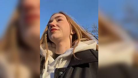 Media: Video of a young woman with long blonde hair, fair skin, and light makeup, wearing a black leather jacket and white scarf, looking up at the clear blue sky, slightly blurred background.