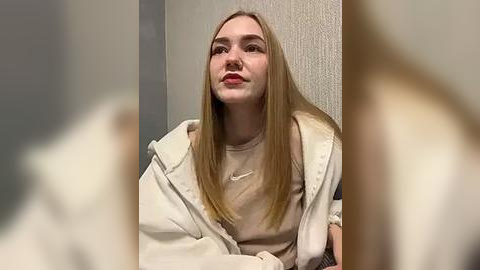 Media: Video of a young Caucasian woman with long, straight blonde hair, wearing a beige Adidas hoodie and a white t-shirt, sitting against a textured beige wall.