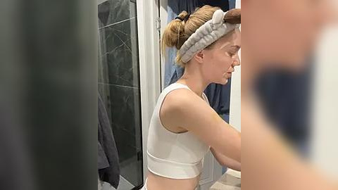 Media: Video of a blonde woman in a white sports bra, wearing a gray headband, applying makeup in a bathroom with a shower and dark towels.