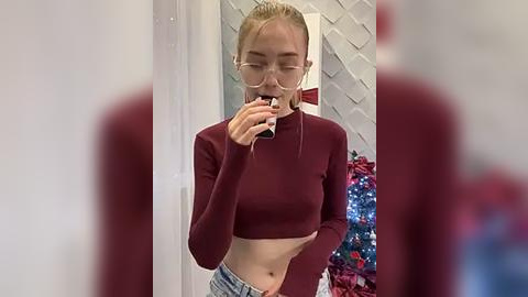 Media: Video of a young woman with fair skin and blonde hair in a maroon crop top, jeans, and glasses, taking a selfie in a modern, festive room with a decorated Christmas tree.