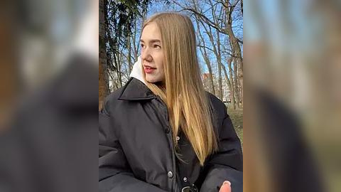 Media: Video of a young woman with long, straight blonde hair, wearing a black winter coat, standing outdoors in a forested area with bare trees and blue sky visible.