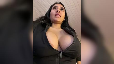 Media: Video of a plus-sized Latina woman with long black hair, wearing a low-cut black top that reveals her large breasts, standing in a dimly lit room with a textured ceiling.