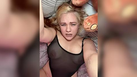 Media: A video of a blonde woman in a sheer black top, lying on a bed with plush toys, her expression one of distress or pain.