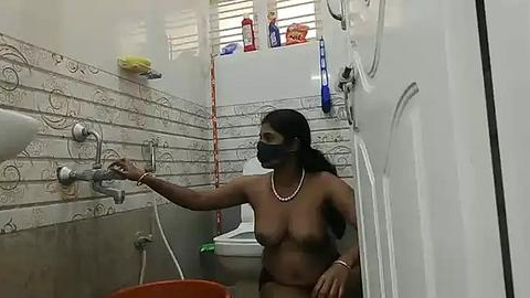 Media: A video of a nude woman with medium breasts and dark skin, wearing a black mask, using a faucet in a small, tiled bathroom with white walls, soap dispenser, and a toilet in the background.