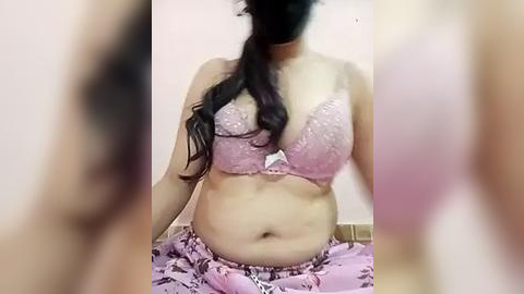 Media: Video of a woman with medium skin tone, wearing a pink lace bra and matching pajama bottoms, sitting on a tiled floor with a blurred background.