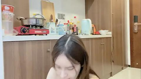 Media: Video of a woman with long, dark hair, sitting on a kitchen floor, surrounded by a cluttered kitchen with a red stove, pans, and various kitchen utensils.