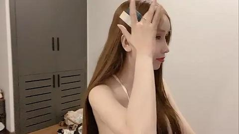 Media: Video of an East Asian woman with long, straight brown hair, wearing a white bra, touching her hair with both hands. She has a pale complexion and is in a simple, minimally decorated room with a gray cabinet in the background.