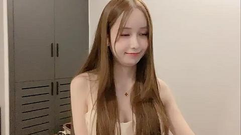 Media: A video of a young woman with long, straight, light brown hair, fair skin, and a slender build. She wears a beige, sleeveless top and a gold necklace. Background includes gray lockers and a plain, light-colored wall.