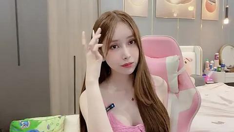 Media: Video of a young Asian woman with long brown hair, wearing a pink lace bra, sitting in a pink gaming chair in a cozy bedroom with pastel-colored walls and soft lighting.