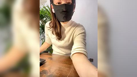 Media: Video of a woman with light skin and long brown hair, wearing a beige sweater, black face mask, and white earbuds, taking a selfie with a blurred background.