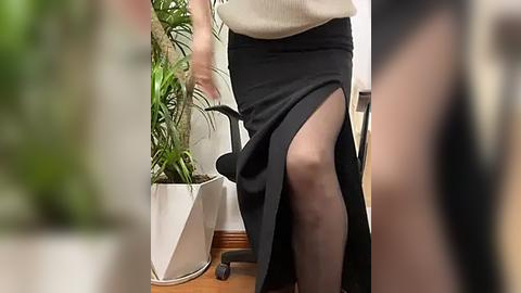 Media: Video of a woman with fair skin, wearing a beige sweater and black high-waisted skirt, with a black high-heeled shoe visible. Background features a white potted plant and office furniture.