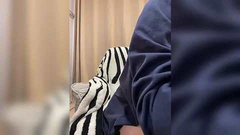 Media: A video of a person in a navy blue sweater, draped in a striped blanket, sitting on a beige sofa with light-colored curtains in the background.