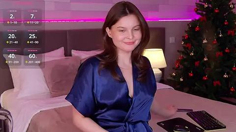 Media: A video of a smiling, fair-skinned woman with long brown hair, wearing a deep blue satin robe, sitting on a bed with a Christmas tree in the background.