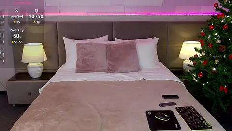 Media: A video of a modern bedroom with a beige bed, two white pillows, a pink throw, a nightstand with a lamp, a smartphone, and a Christmas tree.