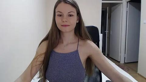 Media: A video of a young woman with long brown hair, fair skin, and a slender physique, wearing a sleeveless, textured gray top, seated indoors with white walls and a black chair in the background.