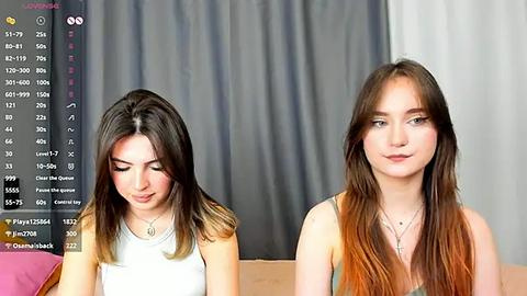 Media: Video of two young women with long brown hair, one in a white tank top, the other in a sleeveless top, sitting side-by-side in a room with gray curtains.