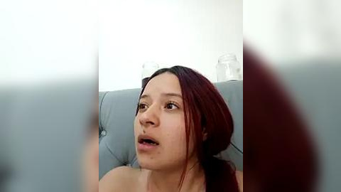 Media: Video of a young Asian woman with straight, shoulder-length dark red hair, light skin, and slightly parted lips, sitting on a grey couch in a brightly lit room.