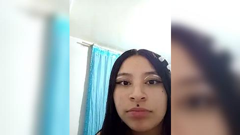 Media: A video of a young woman with straight black hair, light brown skin, and prominent makeup, including dramatic eyeshadow and winged eyeliner. She is in a bathroom with a blue shower curtain and a white wall.