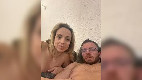 Media: Video of a topless blonde woman with medium breasts lying on a bed next to a bearded man with glasses. Both are smiling.