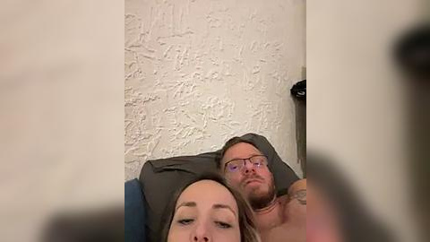 Media: Video of a shirtless man with glasses and a tattooed arm, lying next to a woman with long brown hair, both appearing relaxed on a blue couch against a textured white wall.