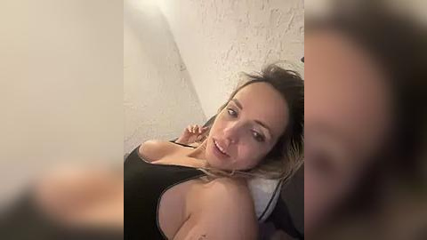 Media: Video of a smiling, light-skinned woman with shoulder-length blonde hair, wearing a black tank top, reclining on a white pillow against a textured white wall.