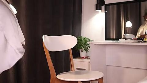 Media: Video of a modern, minimalist bedroom with a white wooden chair, a white desk, dark curtains, a green plant, and a modern black wall lamp.