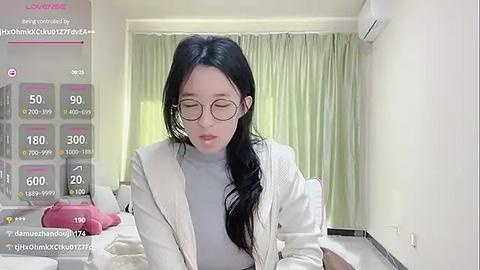 Media: A video of an East Asian woman with long black hair, wearing glasses and a white cardigan, sitting at a table in a modern, light-colored room.